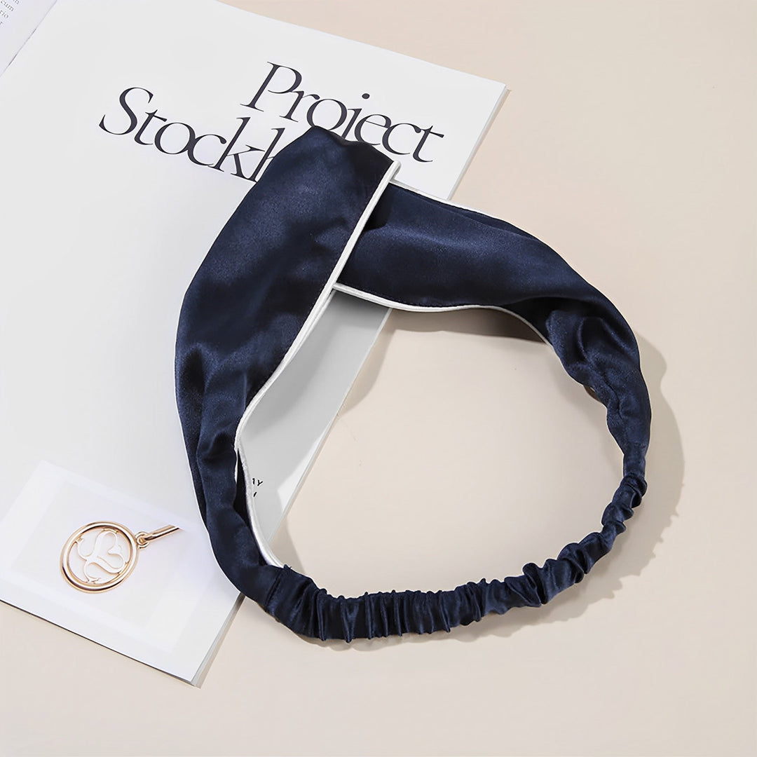 A navy blue Piping Criss Cross Silk Headband by SusanSilk, featuring white trim, is placed on an open magazine next to a gold pendant on a white card.