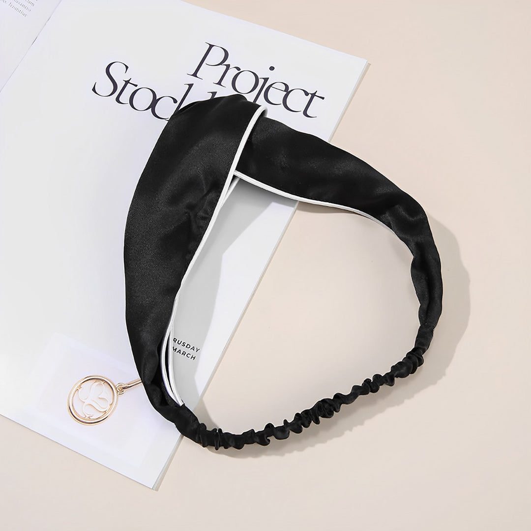 A Piping Criss Cross Silk Headband from SusanSilk, featuring a black satin finish with a white edge detail, is placed on a book titled "Project Stockholm." A partially visible gold pendant accessory nearby enhances the luxurious feel of the SusanSilk fabric.