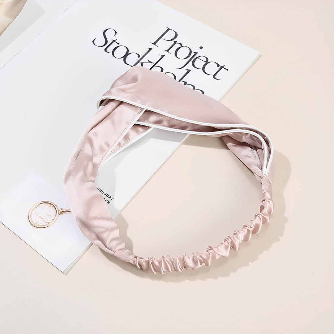 The Piping Criss Cross Silk Headband in pink, designed by SusanSilk and featuring an elastic band, lies atop a magazine titled "Project Stockholm." A white card showcasing the delicate SusanSilk logo, secured by a circular gold clip, is also partially visible.