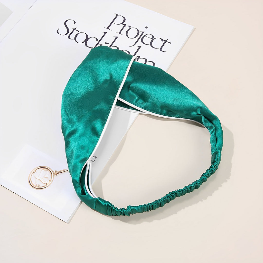 A Piping Criss Cross Silk Headband in green, featuring an elastic band, sits on top of a magazine called "Project Stockholm" along with a circular keychain, subtly evoking the elegance of SusanSilk.