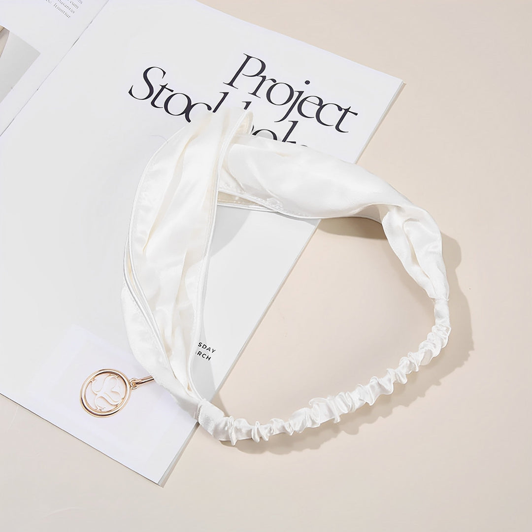 A SusanSilk Piping Criss Cross Silk Headband in white, featuring an elastic band at the back, is placed on top of an open book. A round pendant with an intricate design is also visible. The book, titled "Project Stockholm," seems to complement the luxurious feel of the headband's susansilk fabric.