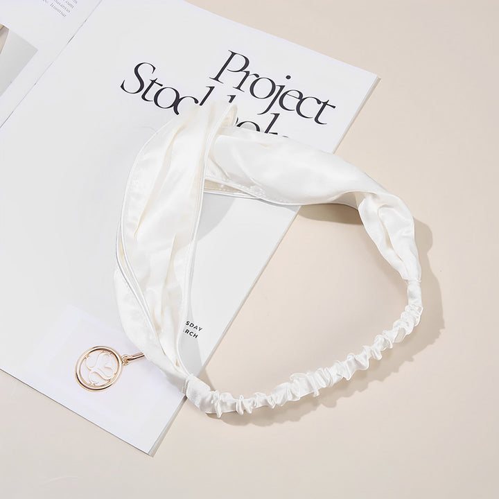 A SusanSilk Piping Criss Cross Silk Headband in white, featuring an elastic band at the back, is placed on top of an open book. A round pendant with an intricate design is also visible. The book, titled "Project Stockholm," seems to complement the luxurious feel of the headband's susansilk fabric.