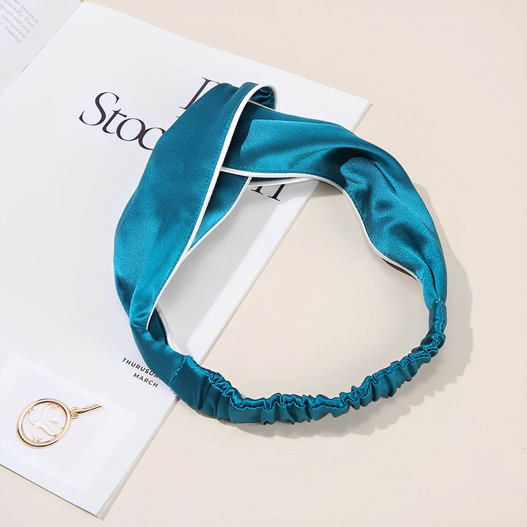 A teal Piping Criss Cross Silk Headband from SusanSilk is placed on an open book next to a gold circular pendant on a beige surface.