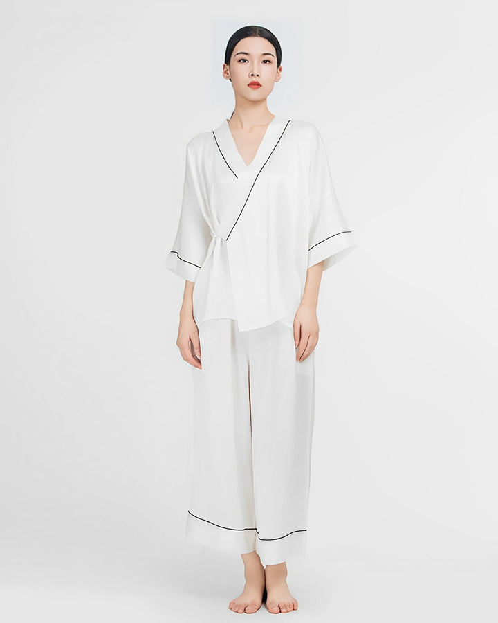 A person stands against a plain white background, elegantly dressed in SusanSilk's 19 Momme Silk Wrap Pajamas Set. The luxurious ensemble features a long-sleeved top and wide-legged pants in white silk with black trim.