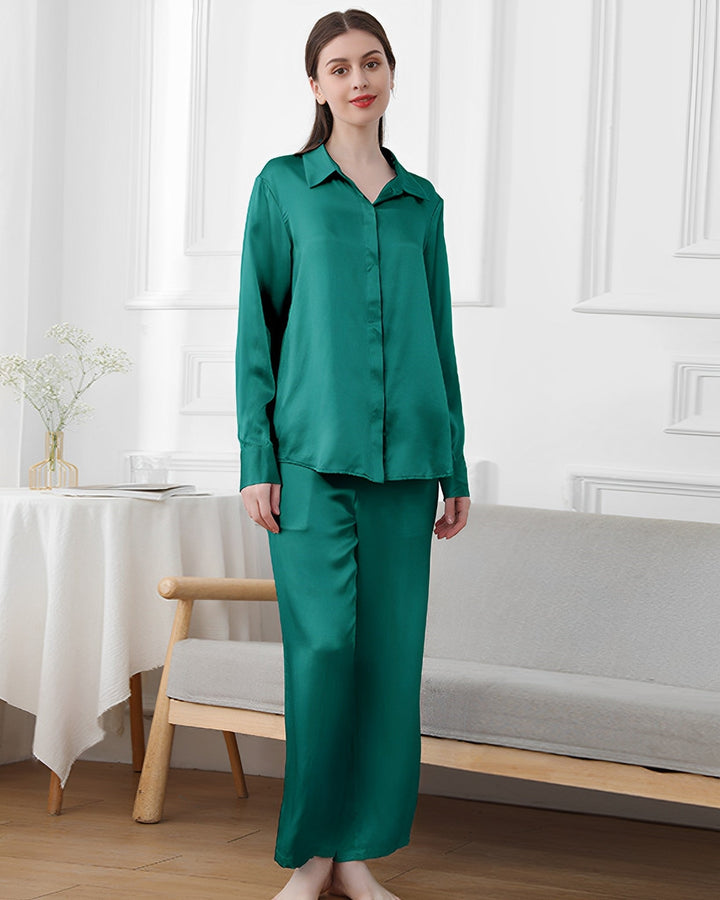 A woman stands indoors wearing SusanSilk's 19 Momme Classic Pajamas Set in green. She is beside a small wooden bench and a round table adorned with a white tablecloth and a vase of flowers.