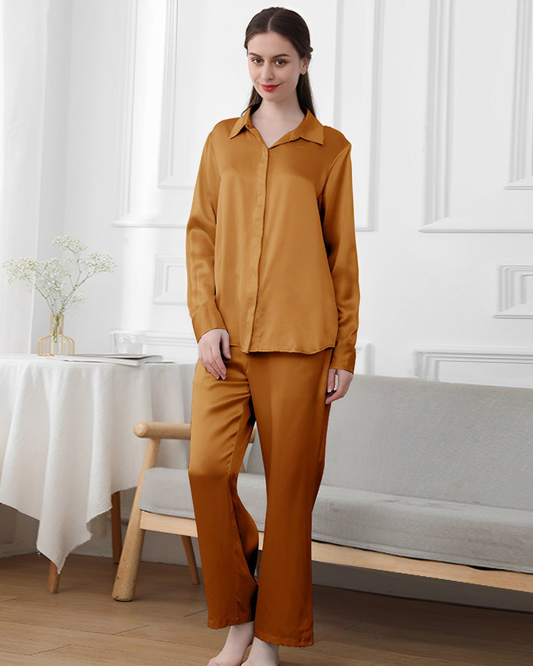 A woman stands indoors wearing SusanSilk's 19 Momme Classic Pajamas Set in a gold color. Behind her is a table with flowers in a vase and a cushioned bench.