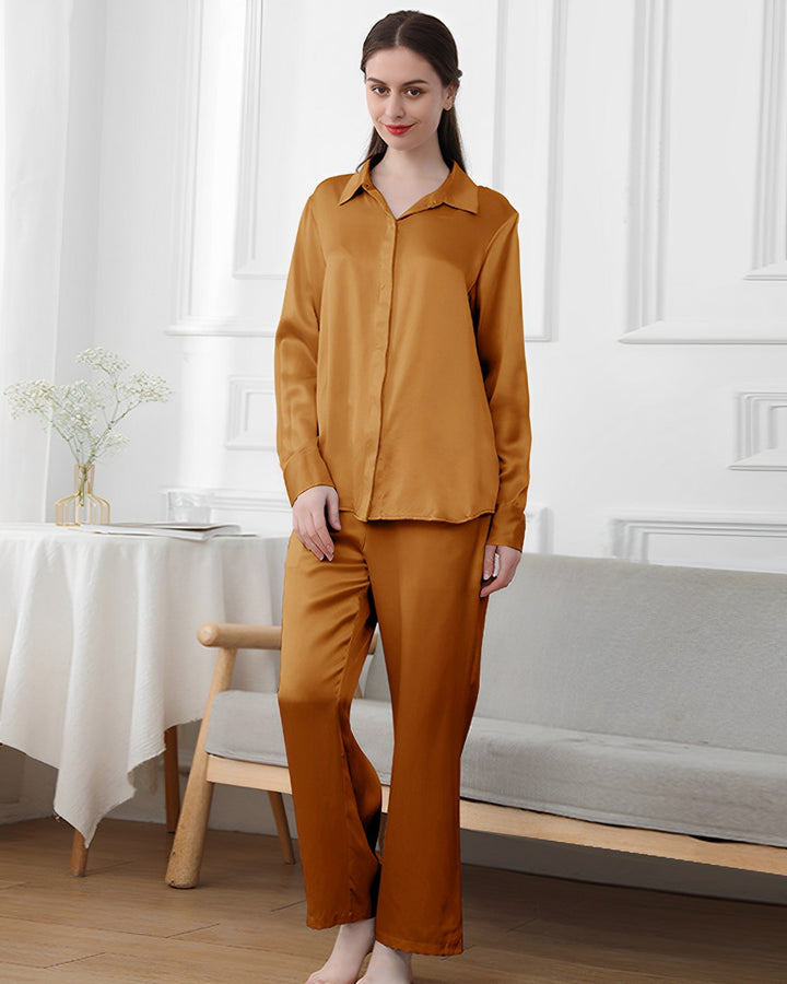 A woman stands indoors wearing SusanSilk's 19 Momme Classic Pajamas Set in a gold color. Behind her is a table with flowers in a vase and a cushioned bench.