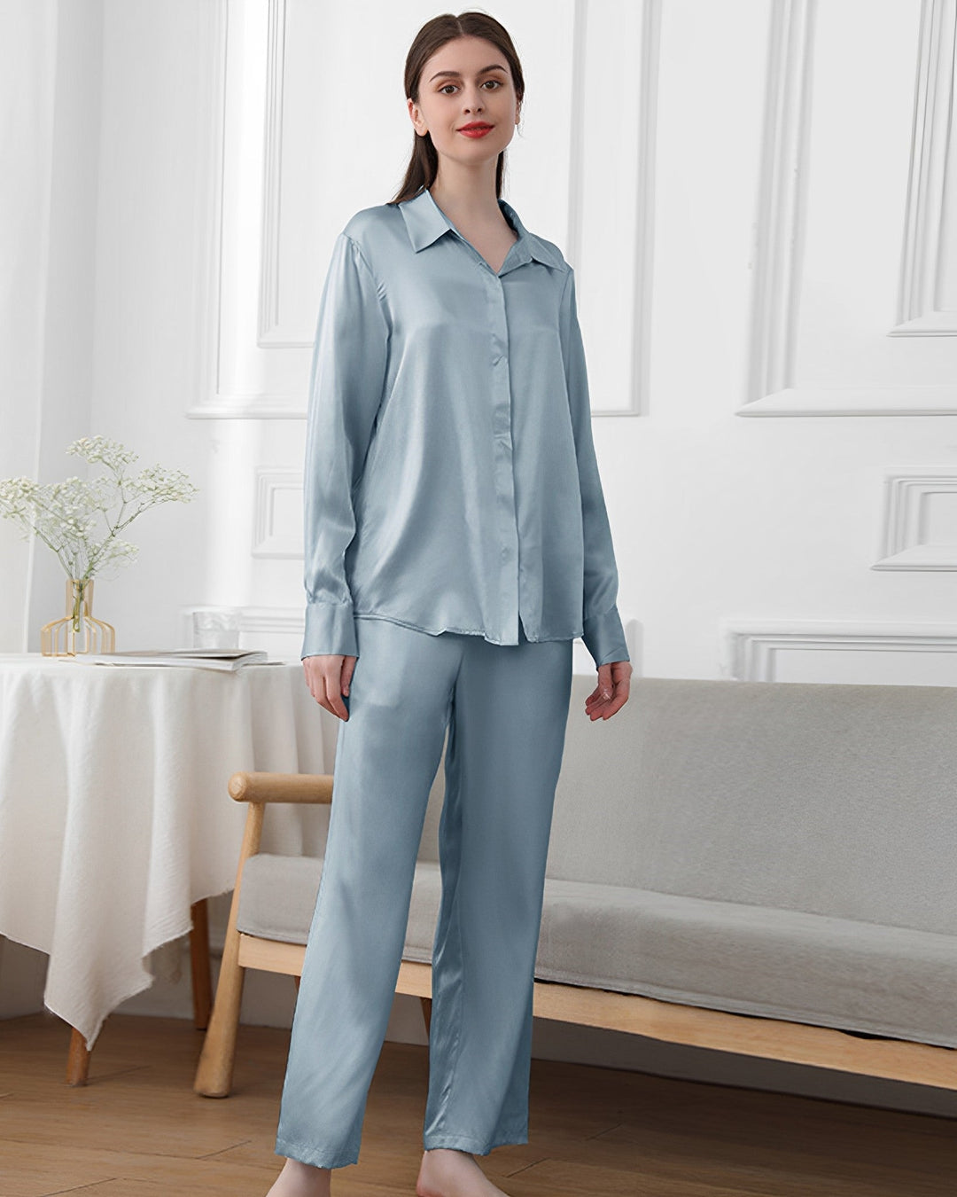 A woman stands in a room wearing a light blue 19 Momme Classic Pajamas Set by SusanSilk. The background features a white wall with molding details, a table with a plant, and a wooden bench with a cushion.