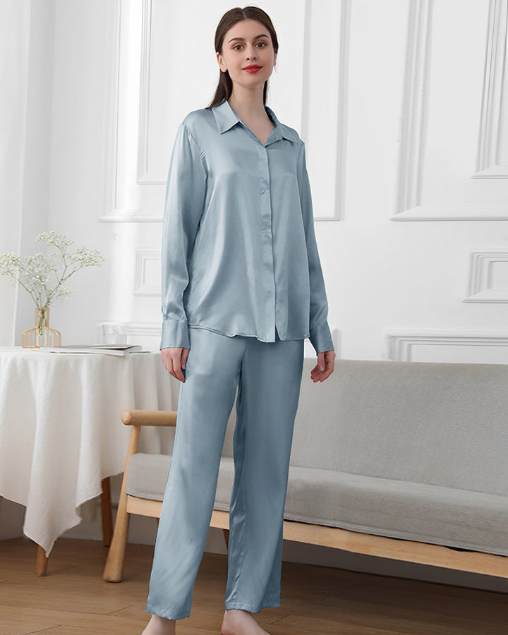 A woman stands in a room wearing a light blue 19 Momme Classic Pajamas Set by SusanSilk. The background features a white wall with molding details, a table with a plant, and a wooden bench with a cushion.