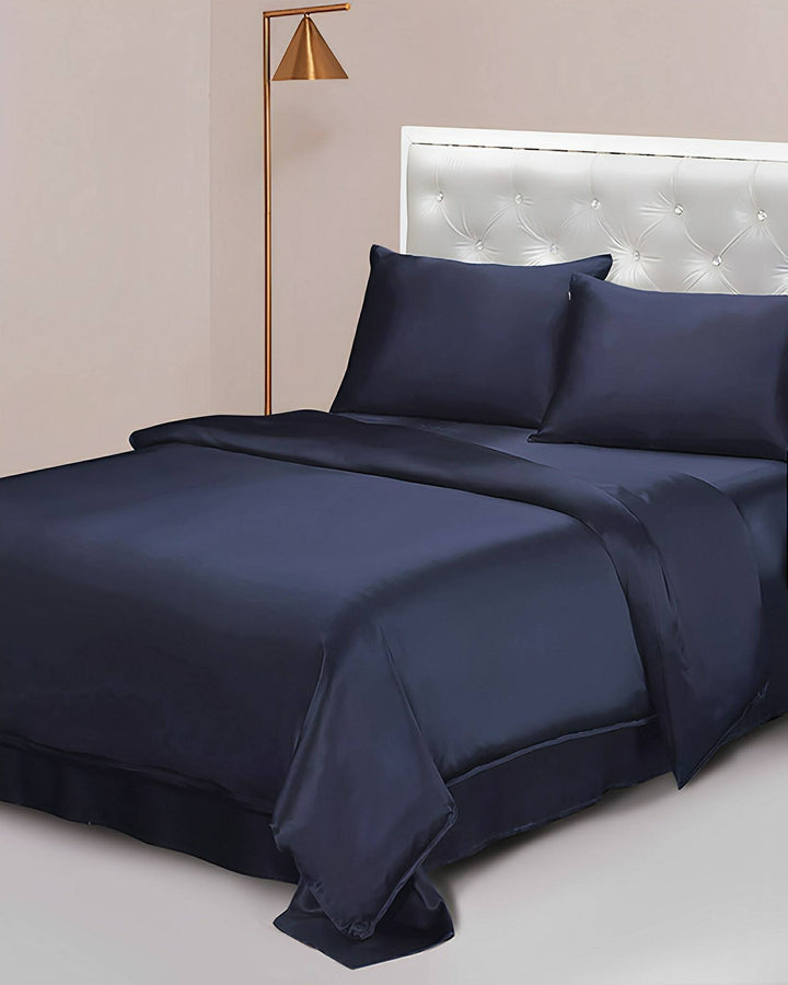 A neatly made bed showcasing a SusanSilk 19 Momme Seamless Silk Duvet Cover in navy blue, complemented by matching sheets and pillows, stands elegantly against a white tufted headboard. A modern gold-colored floor lamp is stylishly positioned next to the bed.