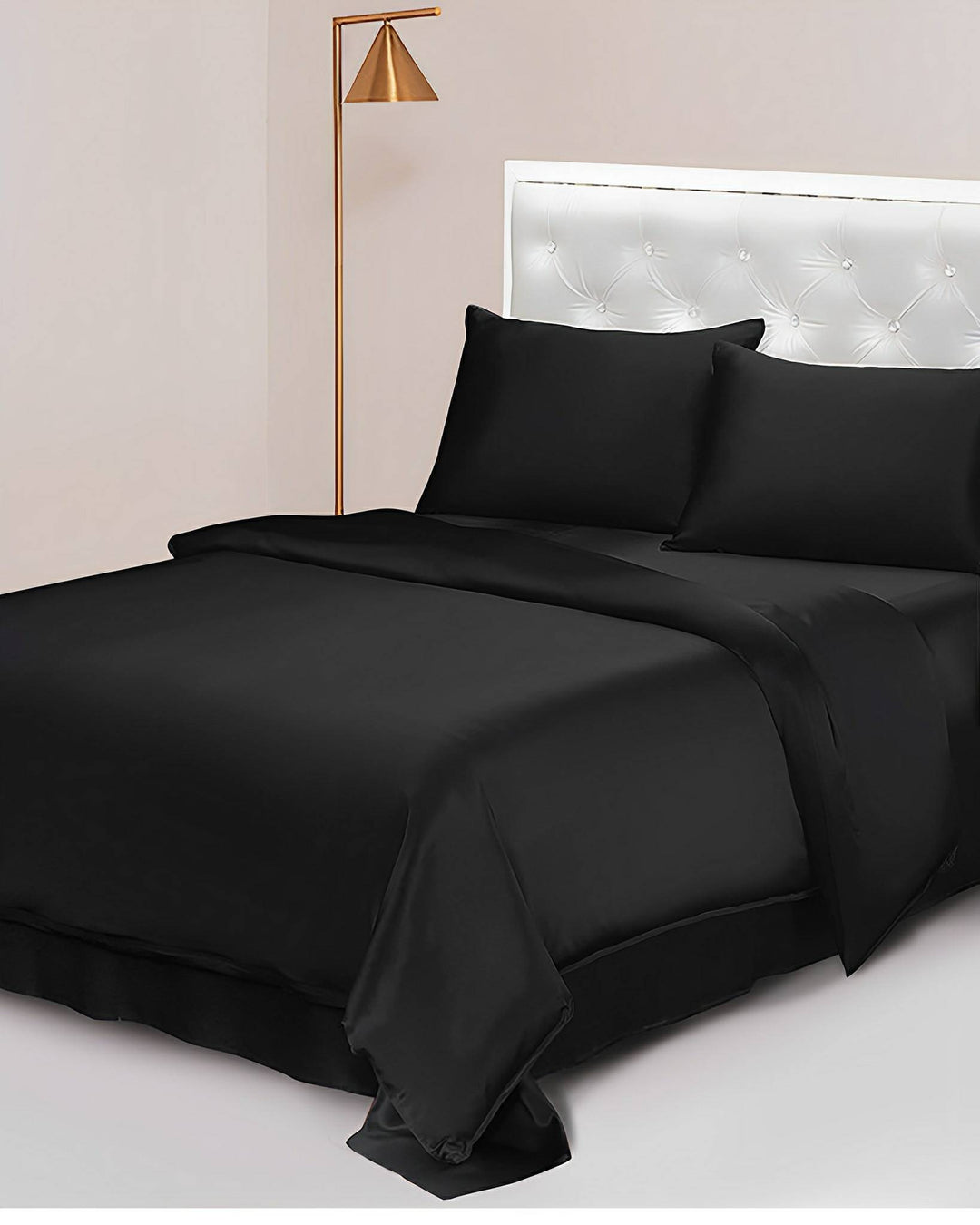 A neatly made bed featuring SusanSilk's 19 Momme Seamless Silk Duvet Cover in black, complemented by matching black silk sheets and pillows. A golden floor lamp stands next to the tufted white wall-mounted headboard.
