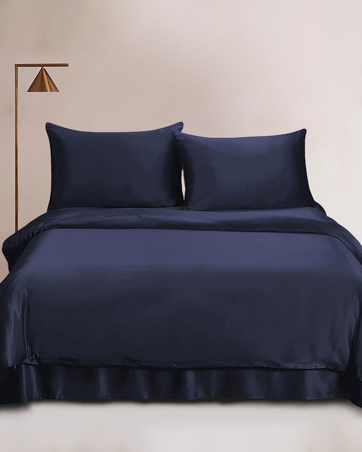 A neatly made bed featuring a 19 Momme Seamless Silk Duvet Cover in dark blue, along with two matching pillows from SusanSilk. A modern floor lamp with a gold finish stands beside the bed, set against a light, neutral-colored background wall.