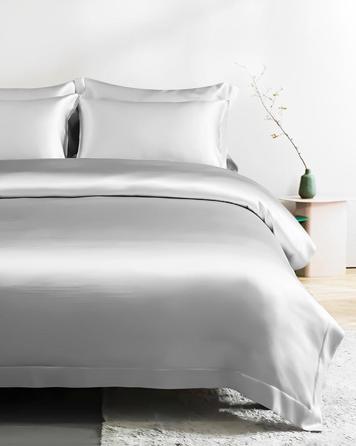A neatly made bed adorned with the 19 Momme Seamless Silk Duvet Cover - Flange Border in light gray from SusanSilk, accompanied by two matching pillows. A small nightstand with a green vase and a plant stands beside the bed, while a light-colored rug partially covers the floor.