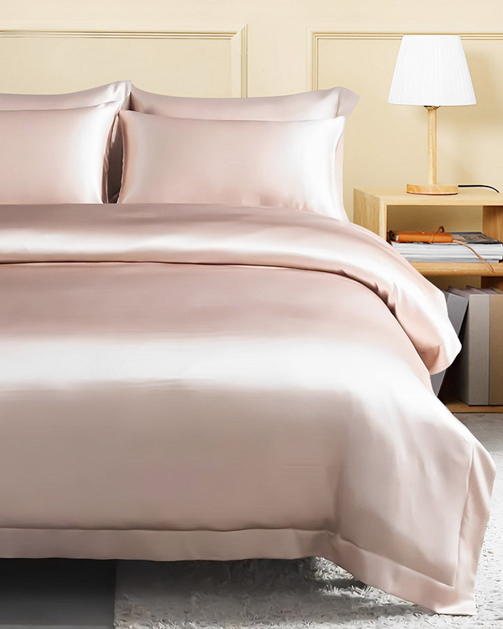 A neatly made bed adorned with a SusanSilk 19 Momme Seamless Silk Duvet Cover with Flange Border in light pink, complemented by matching pillowcases. A wooden nightstand with a lamp and books sits beside the bed.
