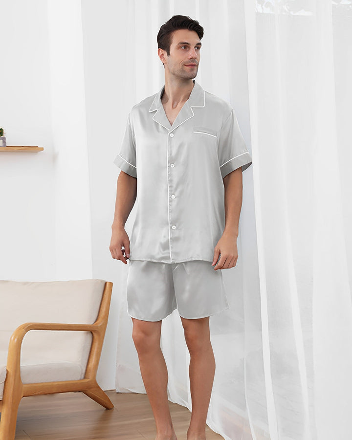 A man stands indoors wearing a light gray short-sleeve 19 Momme Short Silk Pajamas Set from SusanSilk, adorned with white piping. He is near a wooden chair and a sheer window curtain.