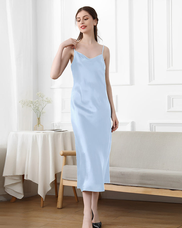 A woman wearing the 19 Momme Silk Sling Dress for Women from SusanSilk stands in a white room, with a bench, small table, and flowers in the background.
