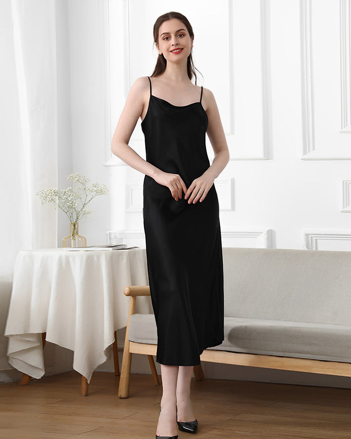 A woman in a sleek, 19 Momme Silk Sling Dress For Women by SusanSilk stands in a white room with minimal decor, including a small table with a tablecloth and a bench. She is smiling and has her hands clasped in front of her.
