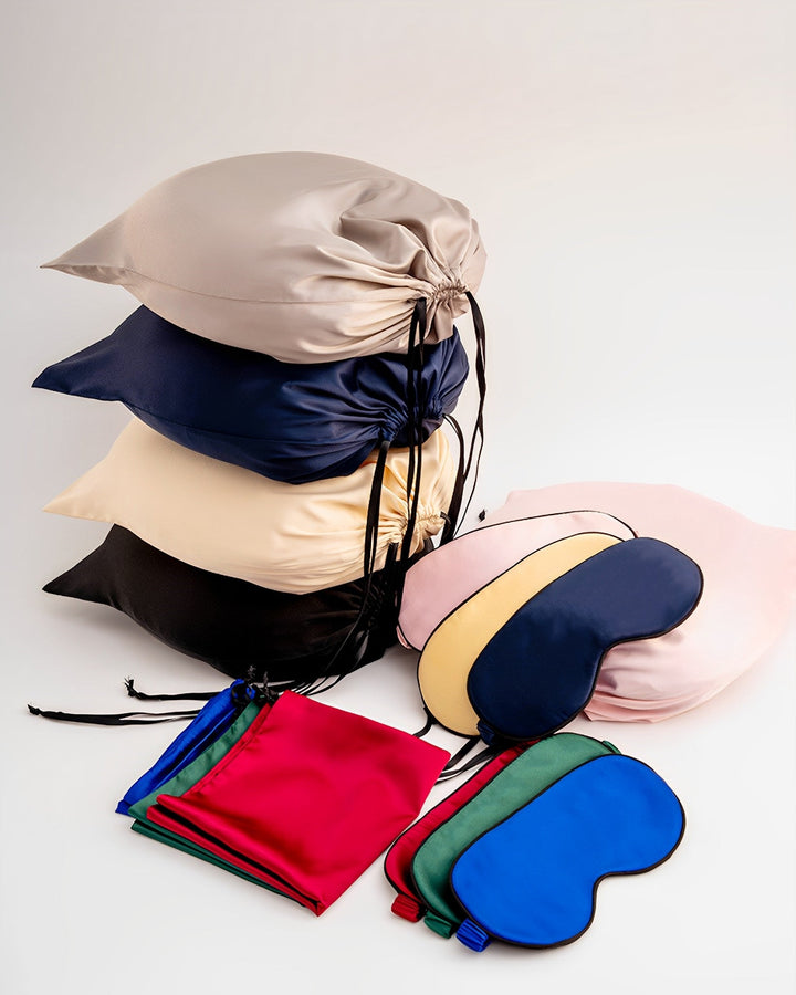A stack of luxurious SusanSilk 19 Momme Silk U-Shaped Pillow Travel Sets in various colors sits beside several folded sleep masks and small drawstring bags, all arranged elegantly on a white background.