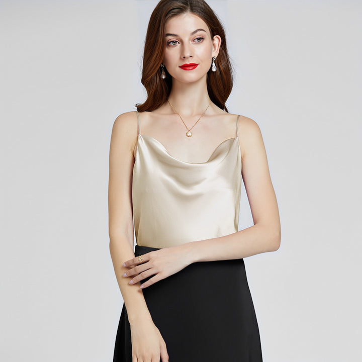 A woman with long hair wears the Elegant Cowl Neck Silk Camisole from SusanSilk, paired with a black skirt. She stands against a neutral background, her hands gracefully positioned together in front of her. Her look is complemented by red lipstick and pearl earrings.