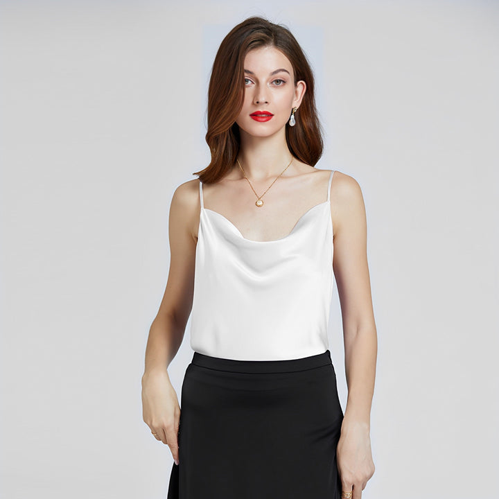 A woman with shoulder-length hair is wearing an Elegant Cowl Neck Silk Camisole by SusanSilk and a black silk skirt, accessorized with a pendant necklace and earrings. She stands against a plain background.