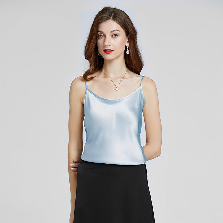 A woman with long hair wearing a light blue Classic Round Neck Silk Camisole by SusanSilk and a black skirt stands against a plain background. She is accessorized with earrings and a necklace.