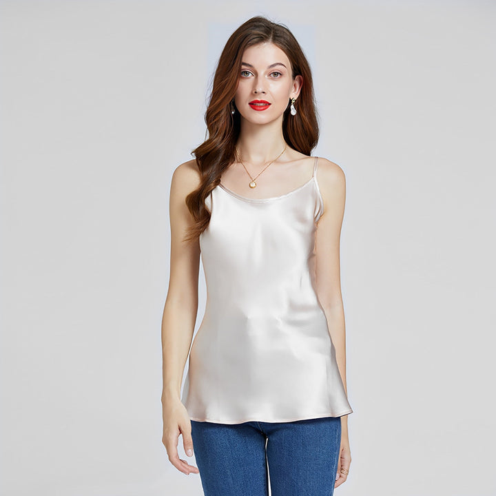 A woman with long hair stands against a plain background, wearing the Classic Round Neck Silk Camisole by SusanSilk and a pair of jeans. She complements her outfit with earrings and a necklace.