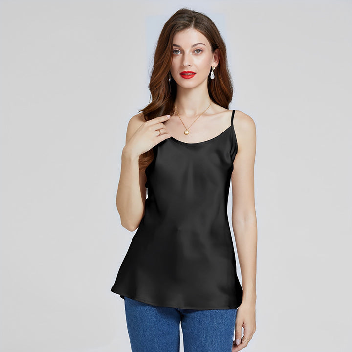 A woman with long hair, wearing the SusanSilk Classic Round Neck Silk Camisole in sleek black and blue jeans, stands against a plain background, looking at the camera.
