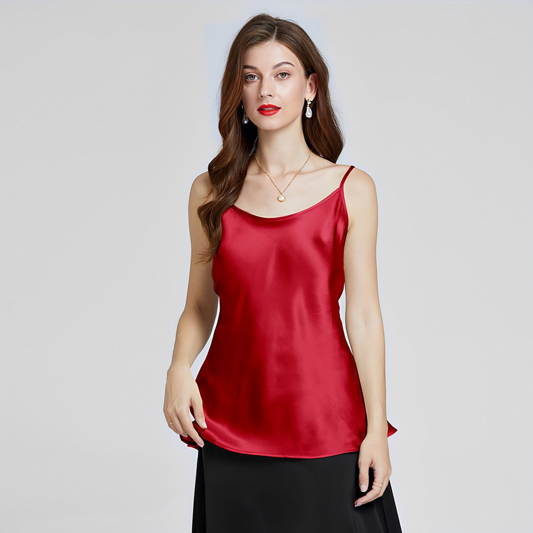 A woman with long brown hair wearing a SusanSilk Classic Round Neck Silk Camisole in red and a black skirt, accessorized with earrings and a necklace, stands against a plain background showcasing her SusanSilk elegance.