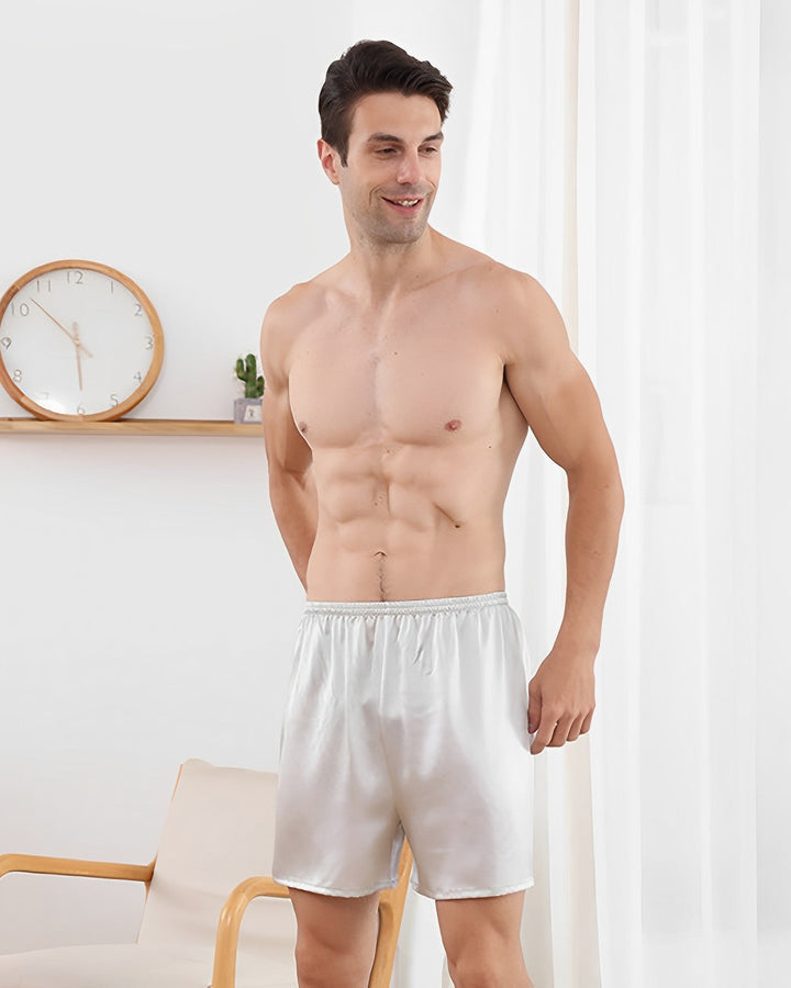 A man stands in a room wearing SusanSilk's 19 Momme Comfortable Silk Boxer, smiling and looking to his right. A clock and small plant are on a shelf in the background.