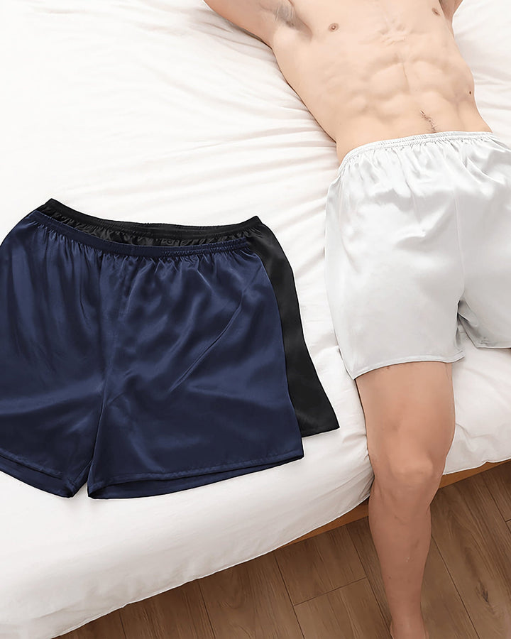 A person is lying on a white bed, partially showing their torso and legs. Two pairs of 19 Momme Comfortable Silk Boxers by SusanSilk, one blue and one white, are placed on the bed.