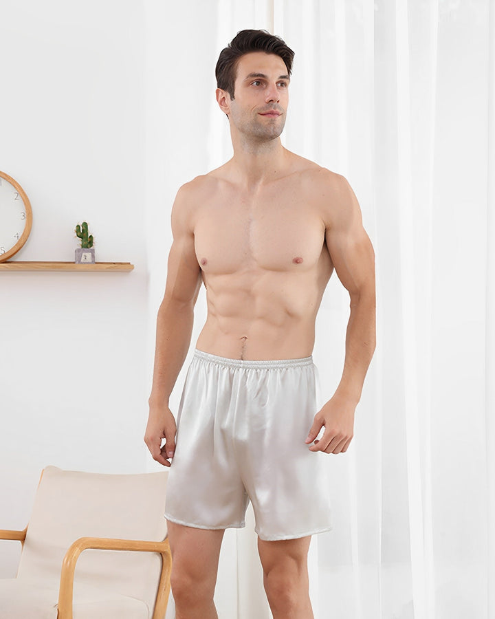A shirtless man wearing 19 Momme Comfortable Silk Boxers from SusanSilk stands indoors near a white curtain and a wooden chair. A wall shelf with a clock and a small potted plant is in the background.