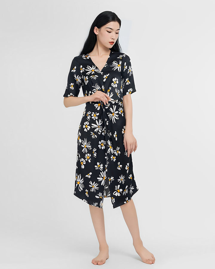 A person stands barefoot on a plain background wearing the Stylish Overlapping Floral Silk Dress by SusanSilk, adorned with white and yellow flowers on a black backdrop, made from luxurious silk.