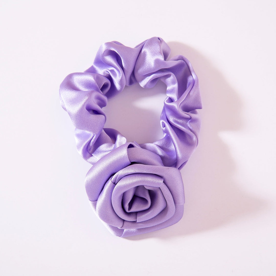 A lavender-colored silk scrunchie with a camellia-shaped fabric detail from SusanSilk, known as the Elegant Camellia Silk Scrunchie.