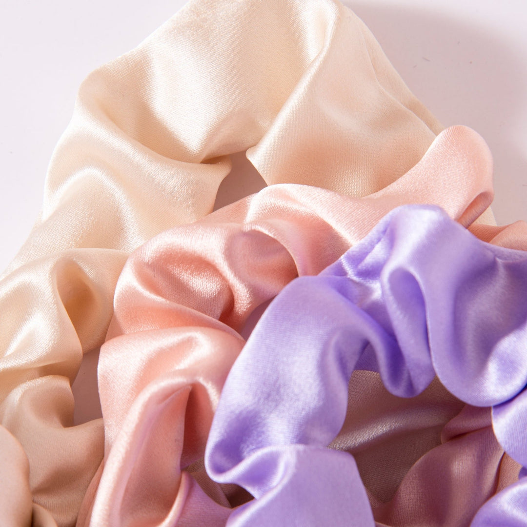 Close-up of three SusanSilk Elegant Camellia Silk Scrunchies in cream, light pink, and lavender colors, arranged overlapping each other.