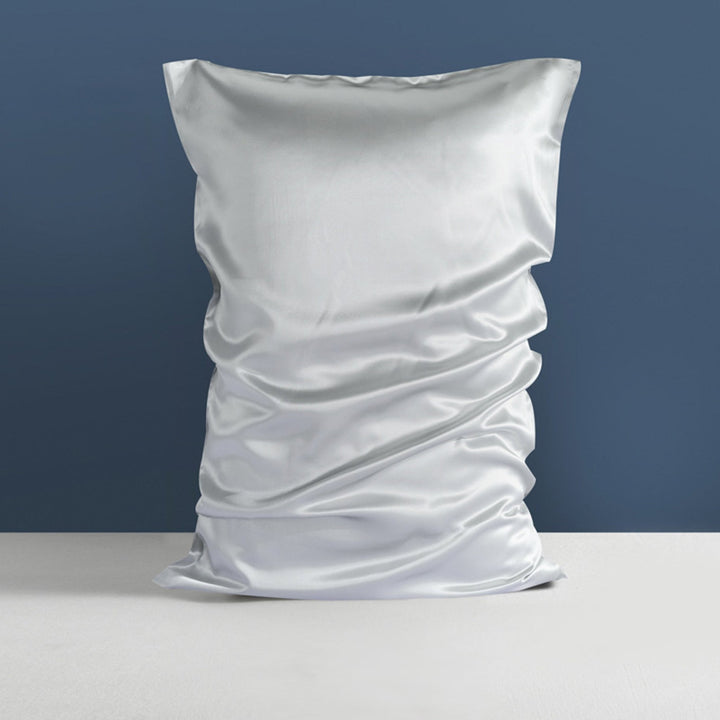 A 22 Momme High Gloss Silk Pillowcase Envelope from SusanSilk, in silver satin, stands against a blue wall on a white surface.