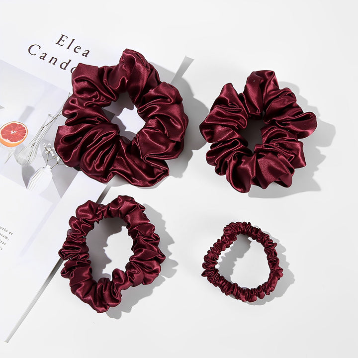 A set of four 22 Momme Luxury Silk Scrunchies by SusanSilk, in elegant burgundy, are displayed on a white surface beside an open magazine.