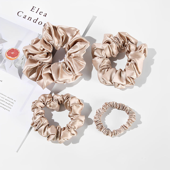 Four scrunchies from the 22 Momme Luxury Silk Scrunchies Set 4-Pack by SusanSilk, come in beige satin and varying sizes, arranged on a white surface next to a partially visible magazine featuring an image of a grapefruit.