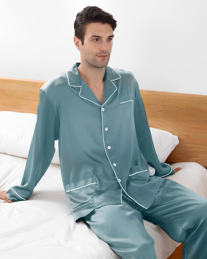 A man in a 22 Momme Lapel Collar Silk Pajamas Set by SusanSilk sits on a bed with white sheets and a wooden headboard, looking forward with a neutral expression.