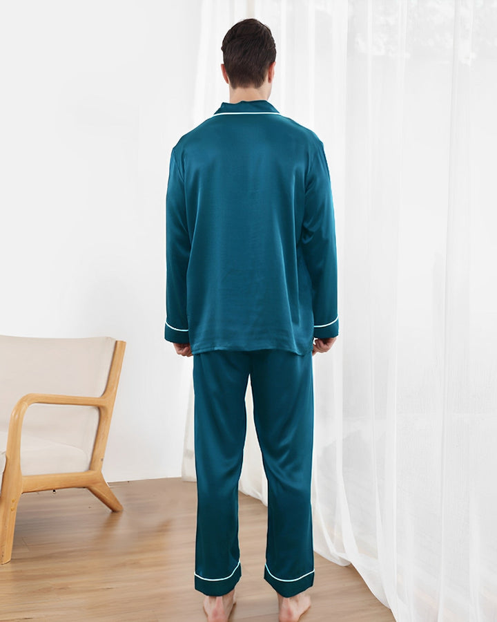 A person wearing a 22 Momme Lapel Collar Silk Pajama Set from SusanSilk in a teal color stands with their back to the camera in a room with a wooden chair and sheer white curtains.