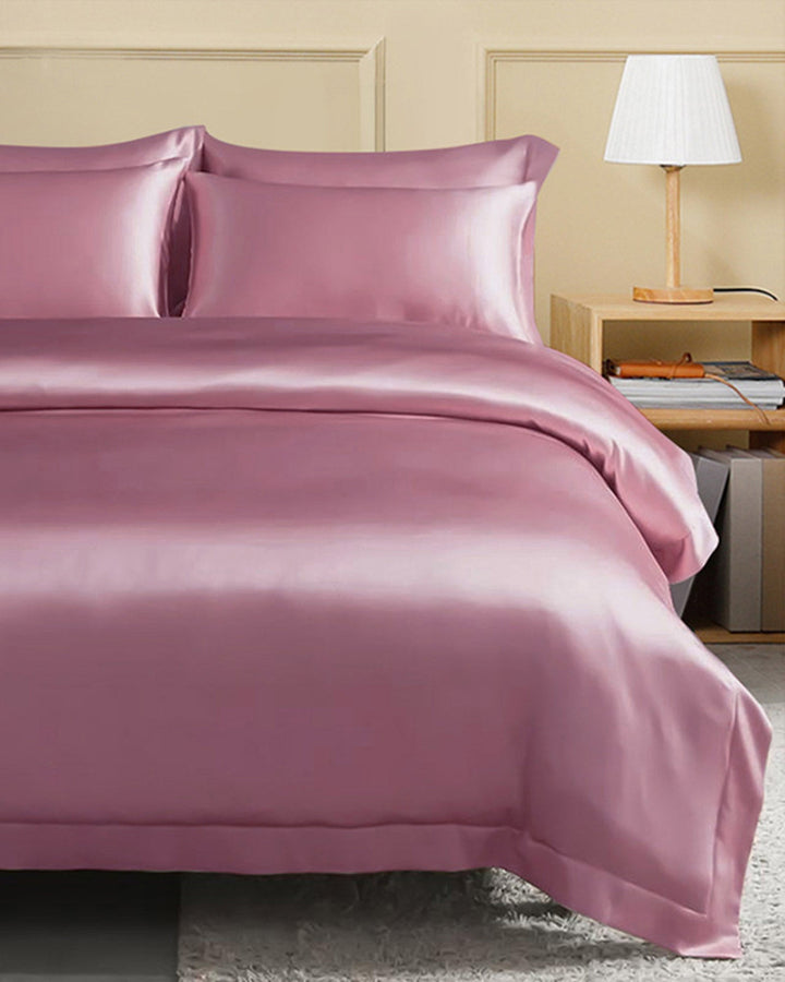 A neatly made bed with SusanSilk’s 22 Momme Seamless Silk Duvet Cover in pink and matching pillows, a wooden nightstand with a white lamp, and books placed on it, all set against a beige wall.