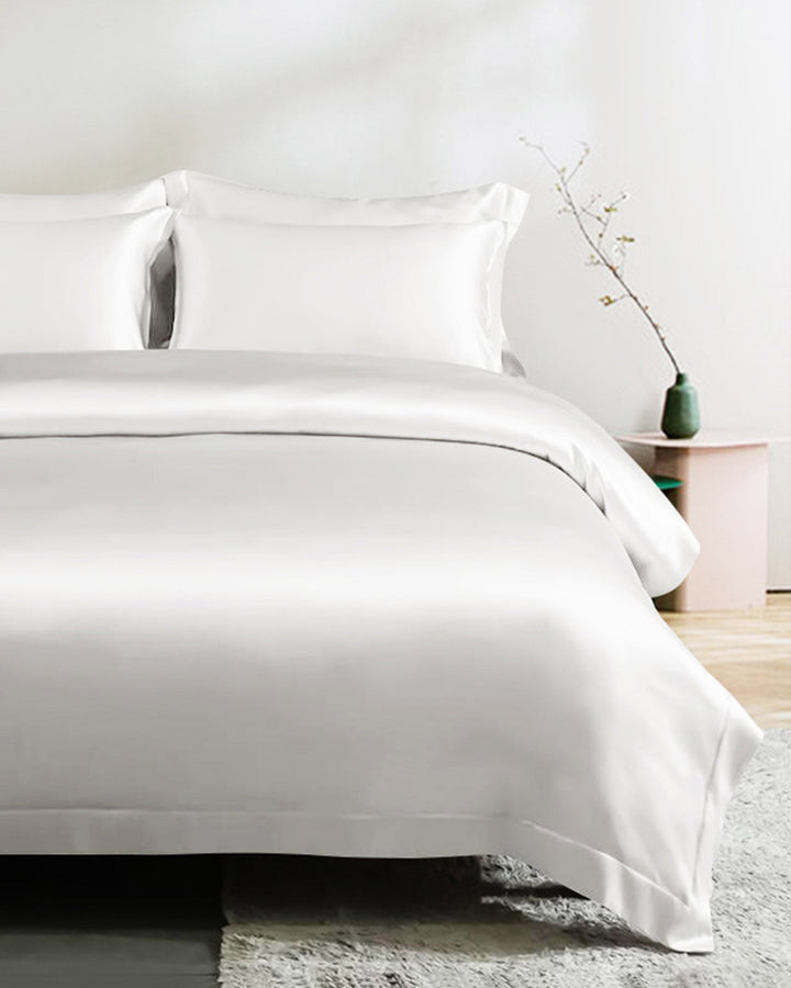 A neatly made bed adorned with the 22 Momme Seamless Silk Duvet Cover - Flange Border by SusanSilk, featuring white silk linens, two pillows, a small side table holding a green vase and a minimalistic plant, set in a bright and modern bedroom.