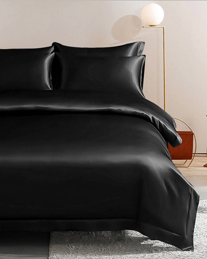 A luxurious black **22 Momme Seamless Silk Duvet Cover - Flange Border** by **SusanSilk** adorns a neatly made bed, complemented by a modern lamp and nightstand in the background.