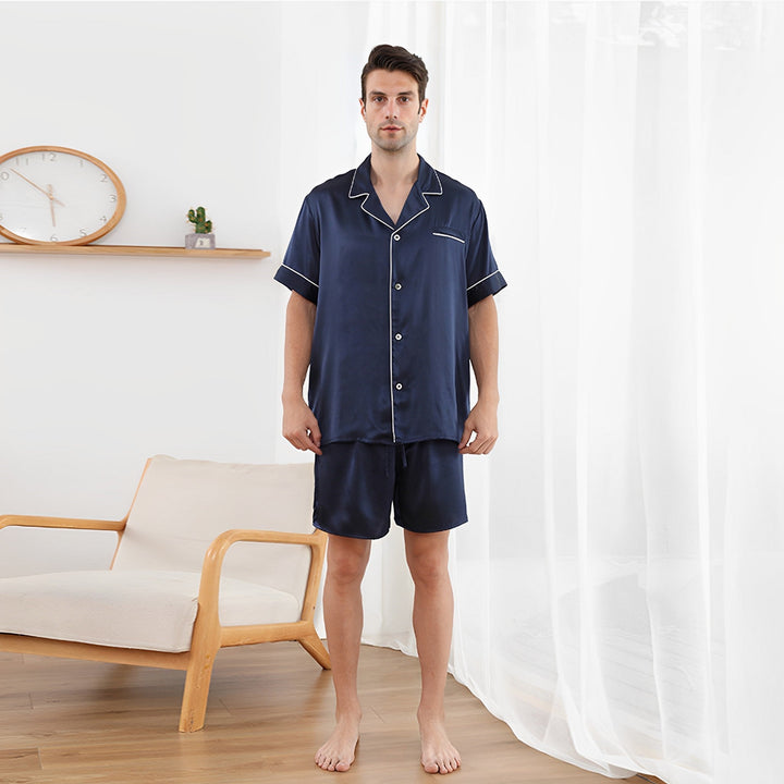 A man wearing a 22 Momme Short Silk Pajamas Set by SusanSilk stands barefoot in a bright room with sheer white curtains, near a modern wooden chair and a wall clock.