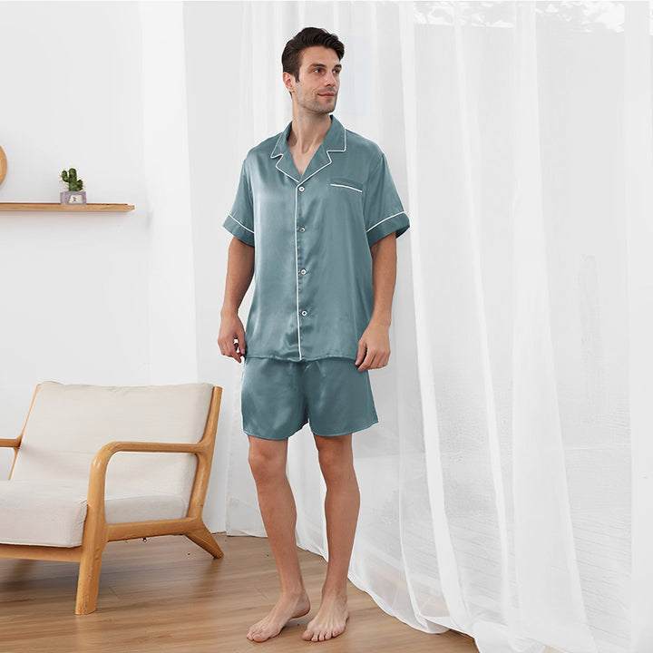 A person stands in a room wearing a matching 22 Momme Short Silk Pajamas Set by SusanSilk, styled in green satin with short sleeves and shorts, next to a beige armchair and white curtains.