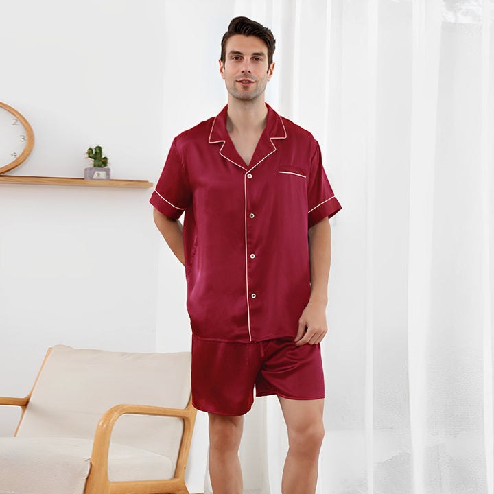 A person stands in a room wearing the 22 Momme Short Silk Pajamas Set from SusanSilk. Behind them is a light-colored armchair and a shelf with a small plant and a clock. Sheer white curtains hang in the background.