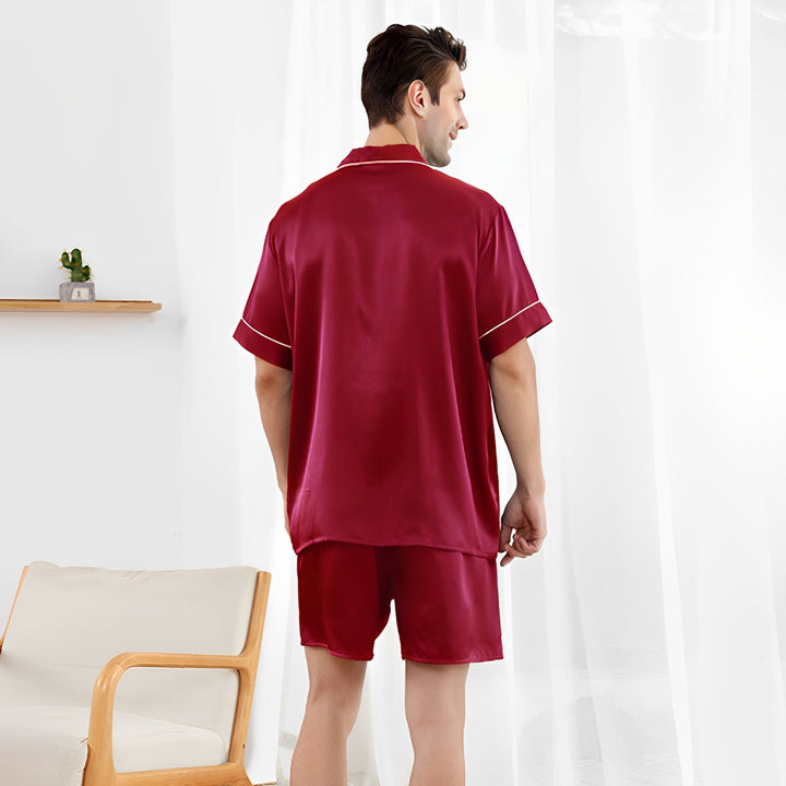 A man stands in front of a window wearing the luxurious 22 Momme Short Silk Pajamas Set by SusanSilk, facing away from the camera. A wooden chair with a cushion is nearby, and a small shelf with a plant adorns the wall. The soft gleam of the silk fabric beautifully contrasts with the cozy room's ambiance.