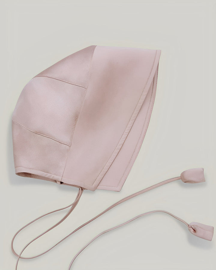 The 22 Momme Silk Sunshade Bonnet Hat by SusanSilk, in beige with stitched panels and a tied chin strap ending in two small fabric weights, is displayed on a light background.