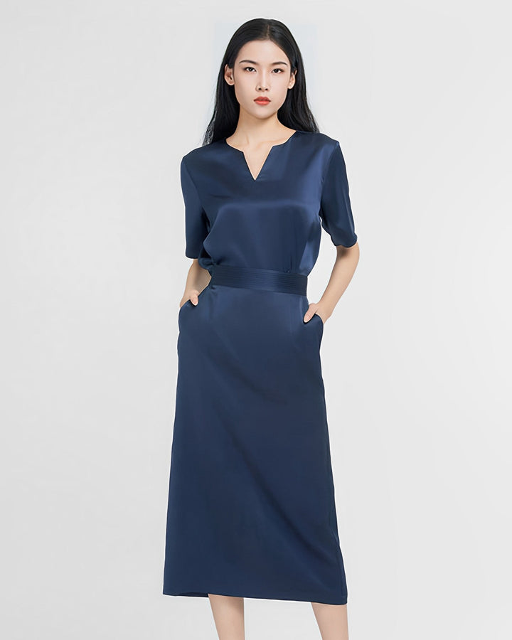 A woman in an Elegant Two-Piece Silk Dress Set by SusanSilk, featuring a dark blue, mid-length design, stands with her hands in her pockets against a plain white background.