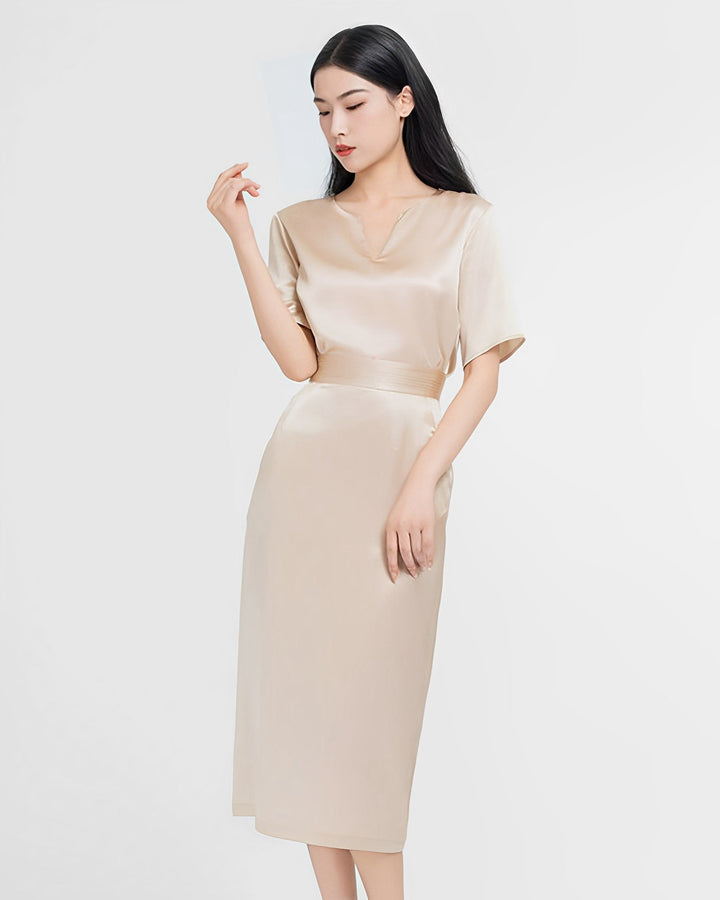 A woman stands against a plain background wearing an Elegant Two-Piece Silk Dress Set in light beige from SusanSilk. The knee-length dress features short sleeves, and she is looking to her left with her right hand raised near her face.