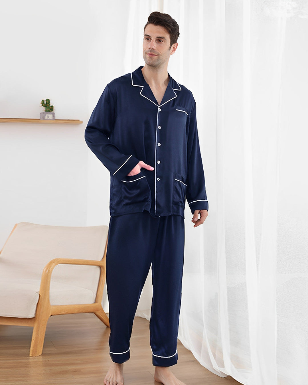 A man stands indoors wearing a SusanSilk 25 Momme Lapel Collar Silk Pajamas Set in navy blue with white piping. He is near a white curtain, a wooden chair, and a shelf with a small cactus.